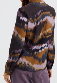 Kvinna iklädd en långärmad blus med abstrakta zigzag-mönster i brunt, svart och lila, tillsammans med lila byxor, visad bakifrån.