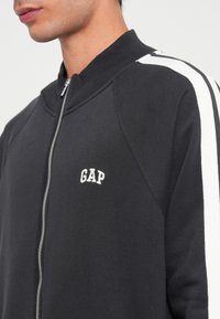 Giacca nera con zip e strisce bianche ai lati, con logo "GAP" in rilievo bianco sul petto. Realizzata in un morbido tessuto misto cotone.