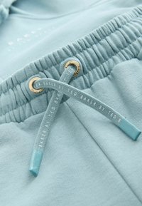 Svetlomodré tepláky v detailnom zábere, zobrazuje elastický pás so zlatými okami a zodpovedajúcimi šnúrkami s nápisom "Ted Baker".