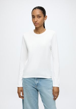 Femme debout devant un fond uni, portant une chemise blanche à manches longues, un jean bleu clair et des boucles d'oreilles dorées, avec une expression du visage neutre.