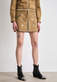 Gonna in suede marrone con chiusura a zip, due tasche con zip, borchie argentate e accenti geometrici. Abbinata a stivali neri alla caviglia con punte argentate.