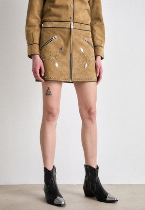 Brauer Wildlederrock mit einem Reißverschluss, zwei Reißverschlusstaschen, silbernen Nieten und geometrischen Akzenten. Kombiniert mit schwarzen Stiefeletten mit silbernen Zehen.