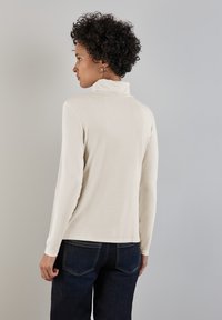 Un haut à manches longues en turtleneck crème, fabriqué en tissu doux, présentant un style ajusté avec un col haut et une légère élasticité, associé à un jean en denim foncé.