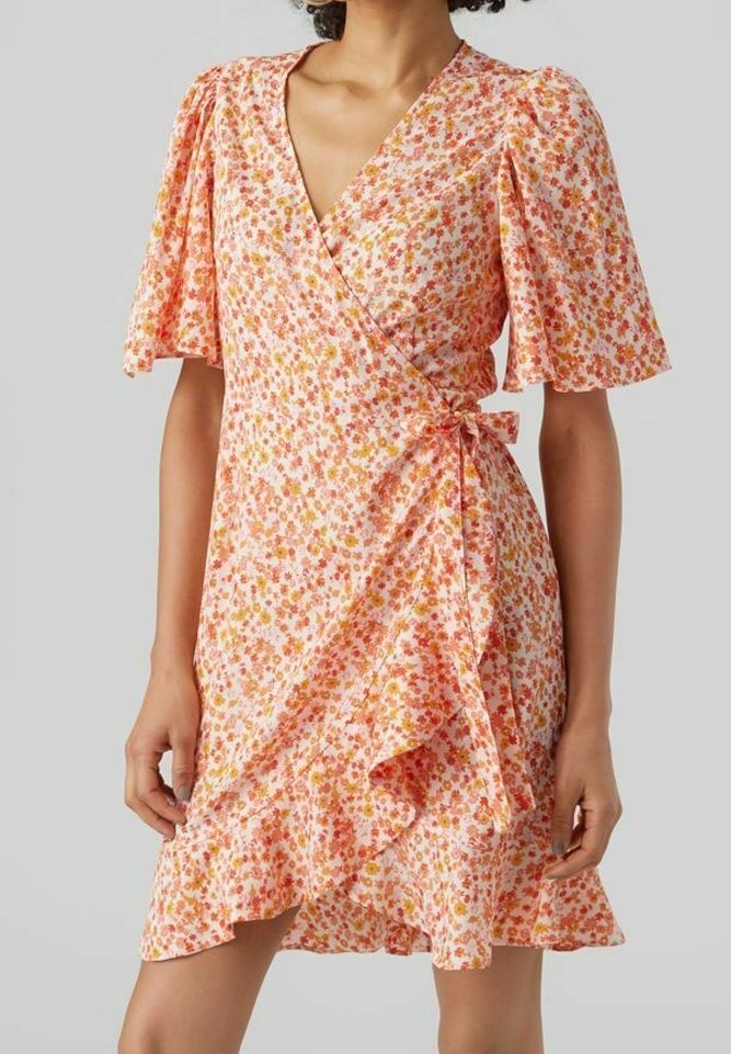 Femme portant une courte robe portefeuille fleurie rose avec des manches volantes et une ceinture à la taille sur un fond uni.