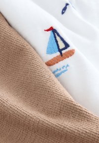 Baumwollstoff, der einen hellbraunen, strukturierten Abschnitt zeigt, der einen weißen Stoff mit einem blau-roten, gestickten Segelboot-Design überlappt.