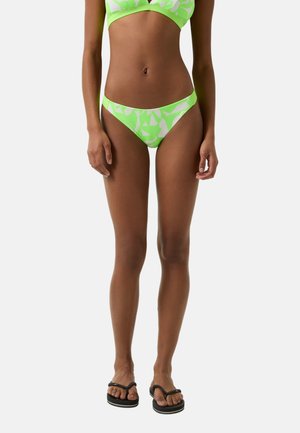 Neon-grünes Bikini-Set aus zwei Teilen mit geometrischen weißen Mustern, bestehend aus einem dreieckigen Oberteil und tief sitzenden Unterteilen. Kombiniert mit schwarzen Flip-Flops.