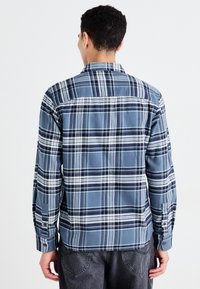 Camisa de cuadros azul con mangas largas, con puños abotonados, cuello trasero y líneas contrastantes en blanco y marino en una tela texturizada.