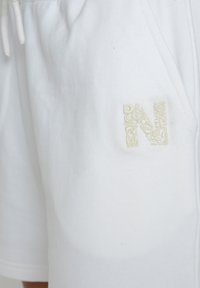 White cotton shorts with a relaxed fit, featuring an embroidered light beige 'N' logo and side pockets. Soft texture, drawstring waistband.