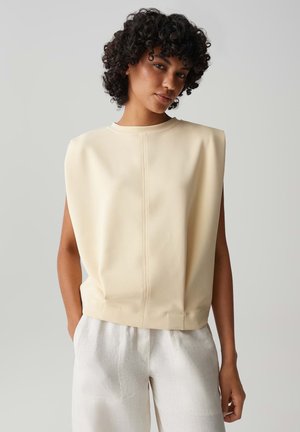 Femme aux cheveux bouclés portant un haut beige sans manches avec des épaules structurées et un pantalon blanc ample, mains dans les poches, fond neutre.