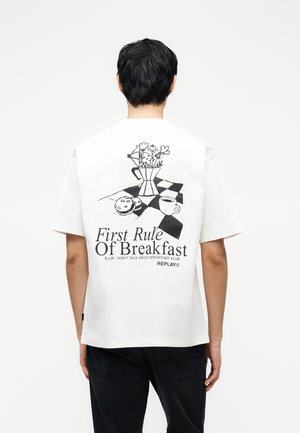 Άνδρας που φοράει λευκό μπλουζάκι με μαύρη εικονογράφηση καφετιέρας, λουλουδιών, μπισκότου, κούπας και κείμενο «Πρώτος κανόνας του πρωινού» στην πλάτη.