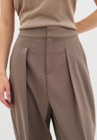Pantaloni a pieghe marroni realizzati in tessuto liscio, con vita alta e tasche laterali, caratterizzati da una vestibilità comoda e un design sartoriale.