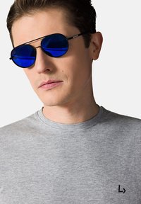 Czarne metalowe okulary przeciwsłoneczne z niebieskimi soczewkami, w kształcie owalnym, odpoczywające na jasnoszarej koszulce z małym czarnym logo na klatce piersiowej.