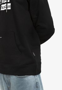 Felpa nera in morbido cotone, con tasca a marsupio e design grafico testuale bianco. Accentuata da un'etichetta 'WASTED' sull'orlo.
