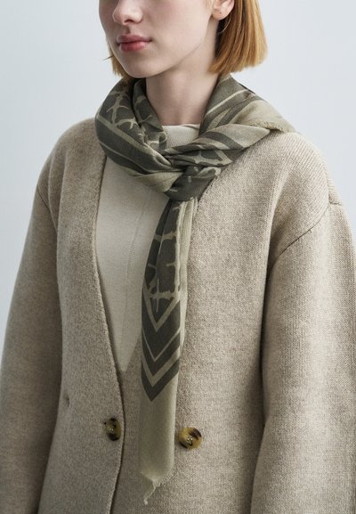 Λαδί πράσινο κασκόλ με γεωμετρικά σχέδια, τυλιγμένο γύρω από το λαιμό, σε συνδυασμό με μπεζ πουλόβερ και υφασμένο ζακέτα.