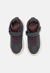 Korkeavartiset tennarit tummansinisestä nahasta viininpunaisilla yksityiskohdilla, joissa on tarranauha, pyöreät nauhat ja valkoiset kumipohjat.