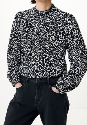 Person trägt eine langärmlige Bluse mit schwarz-weißem Tiermuster, die in hochgeschnittene schwarze Jeans gesteckt ist, mit den Händen in den Taschen.