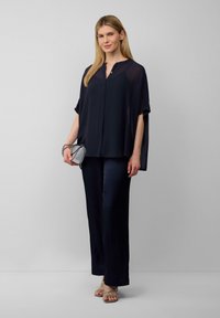 Marineblaue, locker geschnittene Bluse mit kurzen Ärmeln und Knopfleiste, kombiniert mit maßgeschneiderten marineblauen Hosen. Silberne Clutch und helle Sandalen.