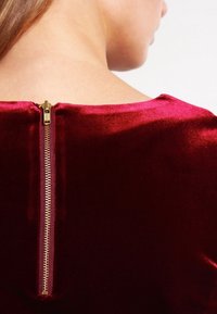 Vêtement en velours bordeaux avec une texture lisse, présentant une fermeture éclair dorée visible au niveau du col au dos, ajoutant un détail fonctionnel.