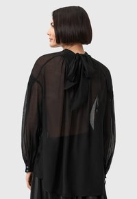 Blusa nera trasparente a maniche lunghe con un laccio sul retro, scollatura arricciata e polsini con bottone. Texture liscia con un design fluido.