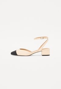 Chaussure à talon ouvert beige en cuir avec un bout noir, design pointu, petit talon carré et une fine sangle de cheville avec une fermeture à boucle.
