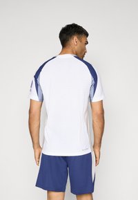 Hvid atletisk T-shirt med blå accenter, korte ærmer og et struktureret mønster. Sammen med marineblå shorts. Fokus på funktionalitet og komfort.