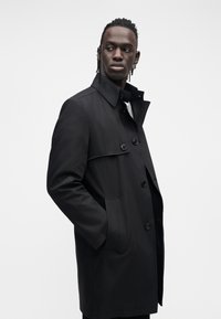 Czarny płaszcz typu trencz o wyraźnej konstrukcji. Posiada wysoki kołnierz, zapięcie na guziki oraz boczne kieszenie. Gładki materiał i dopasowany krój.