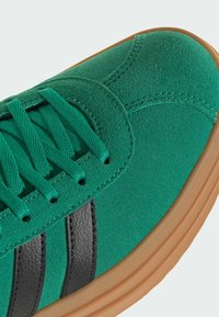 Sneakers en suède vert avec des rayures en cuir noir, présentant une surface texturée, des lacets ronds et une semelle en caoutchouc gum.