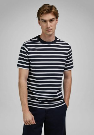 Junger Mann mit hellbraunen Haaren, der ein schwarz-weiß gestreiftes Kurzarmshirt und eine dunkle Hose vor einem grauen Hintergrund trägt.