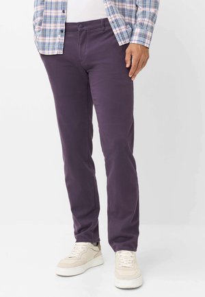 Uomo che indossa pantaloni viola scuro, sneakers beige, maglietta bianca e camicia a quadri blu, in piedi su sfondo bianco.