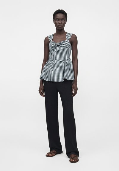 Γυναίκα με μαύρο και λευκό γυναικείο αμάνικο τοπ με σχέδιο γκινγκχαμ και λεπτομέρεια φιόγκου, μαύρες παντελόνες φαρδιές και καφέ σαγιονάρες, στέκεται μπροστά σε απλό φόντο.