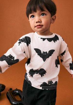 Camicia bianca a maniche lunghe con stampa di pipistrelli neri, scollatura rotonda e vestibilità rilassata, abbinata a pantaloni corti neri. Lo sfondo è di un caldo colore arancione.