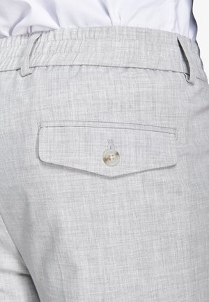 Pantaloni grigio chiaro con tasca posteriore a patta chiusa da un bottone singolo e vita elastica, indossati con una camicia bianca.
