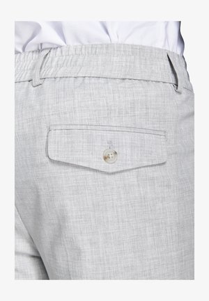 Hellgraue Hose mit einer hinteren Klappentasche, die mit einem einzelnen Knopf gesichert ist, und einem elastischen Bund, getragen mit einem weißen Hemd.