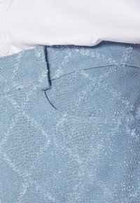 Gros plan d'un tissu bleu clair texturé avec des détails en fil argenté sur un vêtement à côté d'une chemise blanche boutonnée.