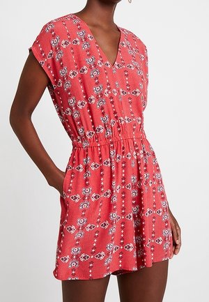 Vrouw draagt een koraalroze korte jurk met bloemenpatroon, V-hals, korte mouwen, elastische taille en zijzakken.