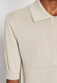 Beige kurzärmeliges Strick-Polohemd mit einem gerippten Ärmelsaum und einer Knopfleiste mit einem Knopf, getragen von einer Person mit mitteldunkler Hautfarbe.