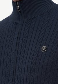 Pull en maille torsadée bleu marine avec un col montant côtelé et un petit badge logo métallique sur la poitrine, fermeture éclair.