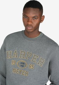 Sudadera gris con texto bordado en amarillo y azul "HARPER 1985 RUGBY TEAM" y "NEYER". Presenta un escote de cuello redondo y puños acanalados.