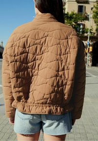 Personne aux cheveux longs et foncés portant une veste matelassée marron et un short en denim bleu clair, debout à l'extérieur par une journée ensoleillée.