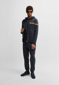 Mörkgrå atletisk träningsdräkt med en zip-up hoodie och joggare. Gyllene strimma och logotypdetaljer; slät textur med en avslappnad passform.