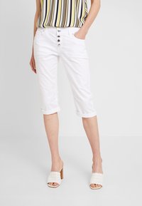 Witte cropped broeken met opgerolde manchetten, voorzien van een knoopsluiting aan de voorkant en zijzakken. Gecombineerd met een gestreepte, mouwloze top en gevlochten muiltjes.