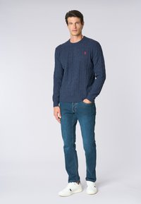Maglione blu navy a lavorazione a trecce con scollatura rotonda e polsini a coste, abbinato a jeans blu slim fit e scarpe da ginnastica bianche. Logo rosso a contrasto sul petto.