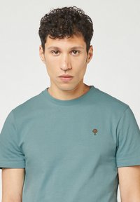 T-shirt in cotone color teal con maniche corte e colletto girocollo. Presenta un piccolo logo di un albero ricamato sul lato sinistro del petto. Tessuto liscio.