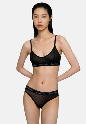 Kobieta w czarnej bieliźnie Calvin Klein z logo na gumce, stoi z lewą ręką za plecami na jednolitym jasnym tle.