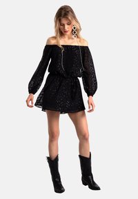 Robe mini noire décolletée sur les épaules avec des manches longues, en tissu texturé et taille cintrée. Associée à des bottes de cowboy noires et des boucles d'oreilles circulaires.