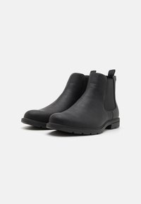 Botas de tobillo de cuero negro con una textura suave, paneles laterales elásticos y lengüetas de tiro. Presentan una punta redondeada y una suela de goma baja.
