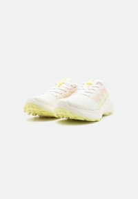 Ζευγάρι λευκά και παστέλ κίτρινα αθλητικά παπούτσια με υφή στην σόλα, στραμμένα προς τα εμπρός σε λευκή επιφάνεια.