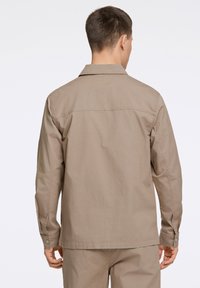 Beige, long-sleeve shirt with a button-up collar, featuring a simple design and textured fabric. Buttons are visible at the cuffs.