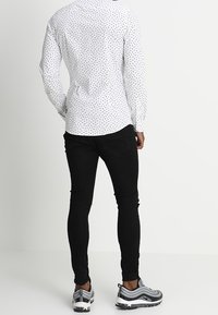 Hombre vistiendo una camisa blanca de manga larga con pequeños patrones negros, jeans ajustados negros y zapatillas deportivas grises, de pie de espaldas.