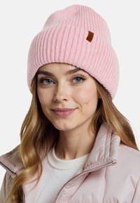 Gorro de punto rosa claro con textura acanalada. Presenta una pequeña etiqueta de cuero marrón. Se lleva con una chaqueta de cremallera rosa claro.